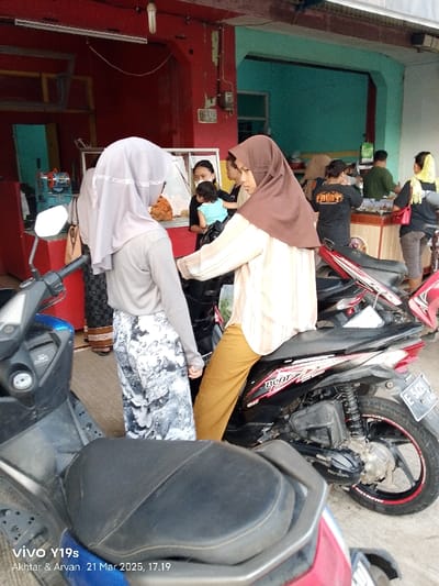 The image shows a street scene in Kecamatan Kroya, Indonesia. A woman in a brown hijab is sitting on a black motorcycle. Another woman in a gray hijab is standing beside the motorcycle, looking at her. A third woman is in the background, wearing a blue shirt and carrying a child. The motorcycle is parked in front of a storefront. The storefront is painted red and has a blue and white awning. There are two other motorcycles parked in the background. There is a sign on the side of the storefront, and the storefront is lit by a lightbulb hanging from the ceiling. The scene is set in a densely populated urban area. The motorcycles are the most prominent subjects in the image, but the women are also significant. The women are the primary subjects, as they are interacting with each other and the motorcycle. The motorcycles are in the foreground of the image and are clearly visible. The storefront is in the background, and the other people are in the background. The image is brightly lit and the details are sharp.