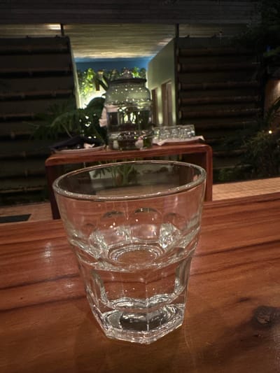 A close-up, sideways shot shows a clear, faceted glass tumbler sitting on a wooden table. The tumbler is empty and reflects the ambient light, highlighting its textured surface. In the background, a dark wooden structure with vertical slats is visible, suggesting an outdoor setting or a room with rustic decor. Beyond this, a dimly lit area with some greenery can be seen, possibly a garden or an outdoor seating area.
The overall scene appears to be in a dimly lit environment, suggesting it might be evening or nighttime. The quality of light hints at artificial lighting rather than natural daylight. The wooden table has a warm, polished finish. The background elements, particularly the bamboo-like structure and foliage, might be indicative of a tropical location. The reflection in the glass and the overall mood lean towards a relaxed, perhaps dining, atmosphere. There is no visible text in the image.
