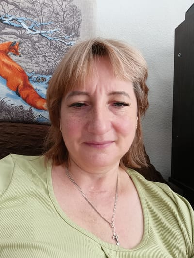 A medium shot selfie shows a middle-aged Caucasian woman with blonde hair and fair skin with freckles. She is wearing a light green ribbed shirt and a silver chain necklace with a cross pendant. Behind her, on the left, is a tapestry depicting a fox in a snowy forest scene. On the right, a dark brown cabinet is visible. The lighting suggests an indoor setting. The woman is looking directly at the camera with a slight smile. There is no specific information or visible clues in the image that indicate the location in the city of Tugulym, Russia.