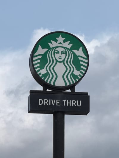 The image shows a Starbucks drive-thru sign against a cloudy sky. The sign features the iconic Starbucks logo, a green circle with a white mermaid holding a tail. The sign is black with white letters and reads "DRIVE THRU." The sign is located in Pocklington, United Kingdom, at the coordinates 53.922, -0.794. The sign is attached to a black pole. The sky is overcast, with white clouds. There are no people present in the image.