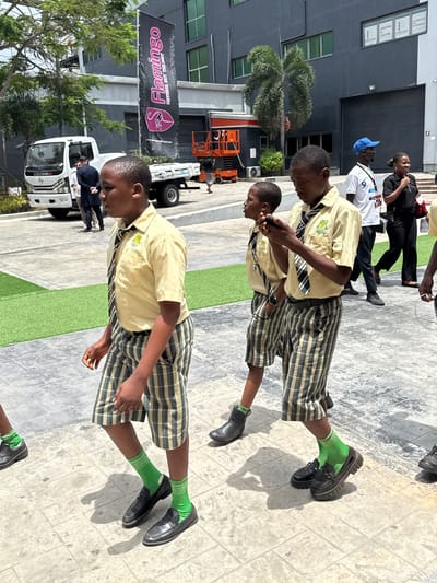 The image depicts a group of uniformed school children standing near a building, possibly in a city. The children are wearing khaki shirts, plaid shorts, green socks and black shoes. One of the children is looking at his phone. There is a banner in the background with the name "Flamingo" and other commercial logos. The presence of a bus, lift and some construction vehicles suggests it might be an area undergoing development. Based on provided text and other details the picture was taken in Itirin, Nigeria.
