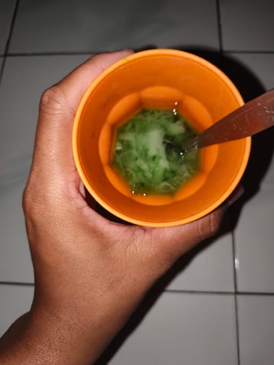 A person's hand holds an orange plastic cup filled with a green, liquid substance and some solid pieces. A metal spoon is partially submerged in the cup. The background appears to be a tiled floor. This image was taken in Barengkrajan, Indonesia.