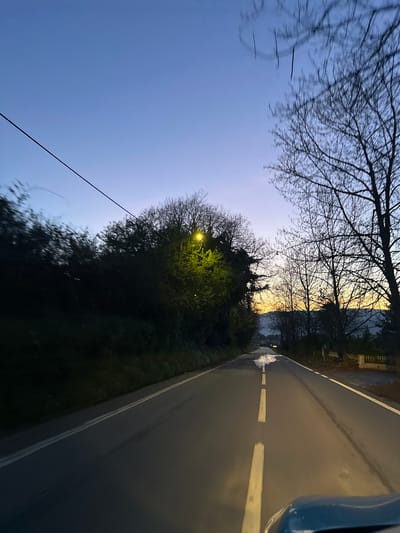 This image displays a two-lane asphalt road in Vila Real, Portugal, under low light conditions consistent with dawn or dusk, where the sky transitions from deep blue overhead to a yellow-orange horizon. The perspective is from inside a vehicle, with a dark blue or teal dashboard or hood partially visible in the lower right foreground. The road features a white dashed center line and solid white shoulder lines. Several wet patches are visible on the road surface, including a prominent, bright, irregular wet area in the middle of the right lane in the mid-distance.
Dense, dark green foliage and trees line the left side of the road, with a bright yellow streetlamp actively illuminating a section of the greenery. A single dark power line extends diagonally across the upper left portion of the sky. On the right side of the road, numerous bare deciduous trees, devoid of leaves, stand silhouetted against the brighter horizon. A low, light-colored stone wall or fence is visible on the right shoulder further down the road. Distant hills are faintly discernible against the horizon. No individuals, animals, or other vehicles are present in the scene.