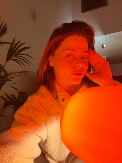 A fair-skinned person with reddish-brown hair, holding their hand to their forehead, looks at the camera. A large, round orange object is positioned near their head, casting an orange hue on their face and surroundings. The person is wearing a white, fuzzy garment. In the background, partially visible, is a potted plant with dark green leaves and a light-colored wall. The lighting suggests an indoor setting.
