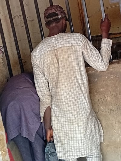 The image, taken in Ayama, Nigeria, depicts two individuals at a barred opening. In the foreground, a male subject is viewed from the rear, wearing a light-colored, faintly plaid long-sleeved tunic. A reddish-brown, textured cap is worn backward on his head, exposing dark hair beneath. His left hand grips a vertical metal bar, part of a series forming a barrier. His right hand holds a black plastic bag positioned at his side. A faint discoloration is visible on the upper back of his tunic. Partially obscured to his left and slightly behind him, a second individual is visible, dressed in dark blue or purple patterned attire, appearing to bend forward. The background consists of a light brown, worn wall. Faded white lettering is discernible on the left side of the wall and the bars. To the right of the main subject's torso, distinct white graffiti reads "17/2/2020 Labiyaffa". The area beyond the vertical bars appears dark.