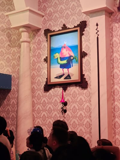The image shows a dimly lit room with patterned wallpaper. A large framed painting hangs on the wall, depicting a pink, anthropomorphic character wearing sunglasses and swim trunks, holding a green inflatable dinosaur. To the left of the painting is a tall, decorative column. To the right is another column with a black, zig-zagging decorative element. In the foreground, several people are visible from behind, their faces mostly obscured, suggesting they are observing the artwork or the room. The overall impression is that of an indoor exhibition or themed space. The image is believed to be from Tongzhou District, China.