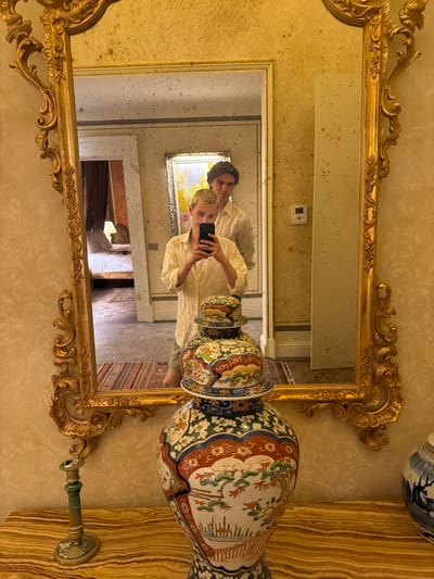 Here is a description of the image:
The image appears to be a photograph taken in the reflection of a large, ornate, gold-colored mirror. The mirror is positioned on a wall with wallpaper featuring a faint, repeating pattern. To the right of the mirror, there's a tall decorative vase with intricate floral and geometric patterns. A candlestick holder is also visible on the right.
The reflection in the mirror reveals two people: one in the foreground holding a smartphone, presumably taking the photograph, and another standing behind and slightly to the side. The people appear to be in a hallway or room with architectural details, including framed artwork, white pillars and a window.
Based on the architectural style and décor evident in the reflection, the image could plausibly be taken inside a historical building or museum in Florence, Italy. However, without additional information, the exact location cannot be definitively determined.