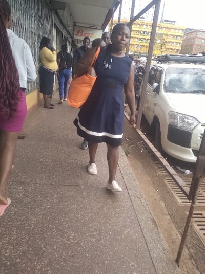 The image, oriented incorrectly and appearing to be rotated 90 degrees clockwise, depicts an outdoor scene on what appears to be a busy urban sidewalk during daylight hours.

In the foreground, a woman stands facing slightly towards the camera, looking directly at it. She is wearing a dark blue, sleeveless dress with white trim along the bottom edge. Her hair is styled in dark braids or dreadlocks. She wears white sneakers and carries a bright orange tote bag slung over her left shoulder, with her right arm extended slightly forward. Her expression is neutral.

Behind and to the left of the main subject, several other individuals are visible, though most are partially obscured or seen from the waist down. One person directly behind the main woman appears to be wearing a dark top. Further back, another person wears blue jeans, and another is seen in a yellow top and a dark skirt. In the very bottom left corner of the frame (which would be the bottom center if correctly oriented), the head and shoulders of a person with long, reddish-brown braided hair are visible, along with glimpses of their legs and feet in what looks like pink footwear. This suggests a queue or a group of people walking together.

The sidewalk itself is made of a light grey, speckled concrete or asphalt, with a visible seam running horizontally across it. To the right of the people (which would be behind them if correctly oriented), a metal railing or fence runs parallel to the sidewalk, separating it from a paved road. A white vehicle, possibly a van or SUV, is partially visible on this road, behind the railing.

In the background, beyond the road and vehicle, several multi-story buildings can be seen, some appearing to be apartments or commercial structures, with facades in shades of yellow and orange. To the immediate left of the main subject, a storefront or building entrance is visible, featuring a roll-down metal security shutter, which appears to be partially closed. There is also a faint, unreadable banner or sign on the wall behind the people, with only a partial word like "Ba..." visible.

The lighting suggests it is daytime, possibly overcast, as there are no strong shadows. The overall scene conveys a typical street environment with pedestrian activity.