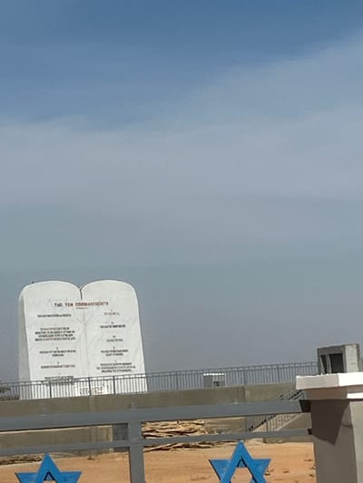 The image depicts a large, white monument shaped like two tablets, inscribed with "THE TEN COMMANDMENTS" and their respective texts in English. This monument is situated outdoors in Kwang, Nigeria, beneath a hazy blue sky. A light grey metal railing runs horizontally across the mid-ground, separating the monument from the foreground. In the immediate foreground, two blue Stars of David are visible, attached to a lower, darker-colored fence or gate structure. The ground beneath and around these elements appears dry and sandy, with some brown, possibly wooden, material visible on the ground in the mid-ground. A partial concrete structure is visible on the right side of the foreground. No people are present in the scene.