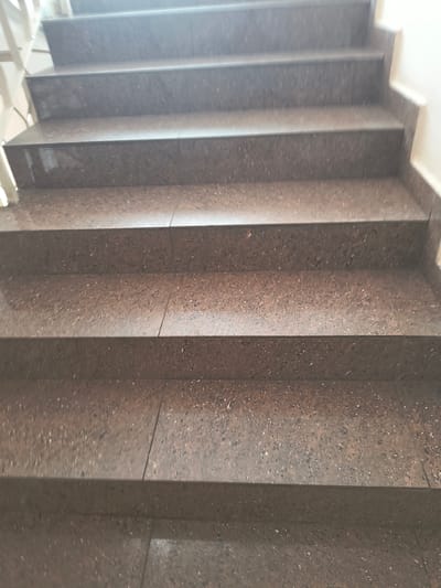 The image shows a close-up view of a flight of granite stairs in an indoor environment. The stairs are made of speckled reddish-brown and grey granite tiles, with visible grout lines. The perspective is from a low angle, looking up the stairs. The lighting appears to be artificial, casting shadows on the steps. There are no people or other objects visible on the stairs. The overall impression is one of a utilitarian and well-maintained public staircase. The location in Rivne, Ukraine, is not evident from the visual information provided.