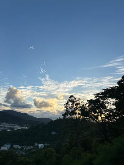 This is a vertically oriented, outdoor, daytime photograph taken in Ratsada, Thailand. The scene depicts a landscape view with a gradient of blue sky transitioning into mountainous terrain densely covered in green foliage and dotted with white buildings. The sky is filled with a mixture of thin, wispy clouds and larger, fluffier cumulus clouds. Some of the clouds are illuminated by the sun, giving them a warm, golden hue, suggesting it is either sunrise or sunset. Silhouetted against the sky and the illuminated clouds are the dark green leaves and branches of trees in the foreground, framing the view. The mountains in the background appear dark and rugged, partially obscured by trees. White structures, likely houses or buildings, are visible nestled on the slopes of the mountains, suggesting a residential area on the hillside. The overall impression is one of natural beauty with a touch of human habitation. There is no visible text in the image.