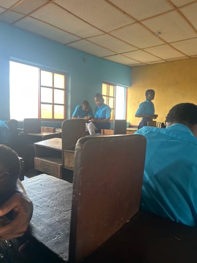 The image depicts an indoor scene within a classroom in Ikorodu, Nigeria, shot from a low perspective, looking towards the front or side of the room. The photograph is oriented sideways, but when rotated, it shows several students, primarily young people, dressed in light blue uniforms, seated at wooden desks and benches.
In the immediate foreground, part of a student's head and hand are visible, seated at a dark, worn wooden desk. Beyond this, rows of similar wooden desks and attached benches extend into the room. Bright sunlight streams intensely through large, multi-paned windows on the left side of the frame, creating significant glare. The walls of the classroom are painted in two distinct colors: a light blue where the windows are situated, and a warm yellow on the opposite wall. The ceiling features a grid pattern, possibly constructed from light-colored panels with wooden supports, and a single light bulb is faintly visible.
Further back in the classroom, one student is seen standing or leaning near their desk, appearing to hold a small object, perhaps a phone or book. Other students are seated in various positions, some looking forward. The overall activity suggests a routine school day, with students either attending a lesson, studying, or waiting. The strong sunlight indicates it is daytime. The setting appears modest but functional, typical of a local school environment. No discernible text is visible in the image.