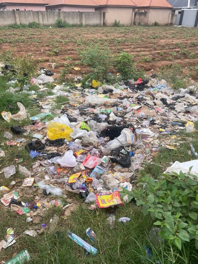 The image depicts a scene of significant litter accumulation on the ground, bordering a patch of tilled earth with sparse green vegetation. The litter consists of a wide variety of discarded items, including plastic bags, wrappers, bottles, and other unidentifiable debris. Some of the plastic bags are white, black, and colorful, while others appear to be clear or translucent. Among the trash, a yellow packet with the text "Indomie" is clearly visible, suggesting it is a food product wrapper. Another item with blue and white text and images is also present. The ground itself is a mix of dry earth and sparse grass, with more dense green foliage visible on the right side of the frame. In the background to the left, part of a building with a terracotta-colored roof and a concrete wall is visible, indicating an urban or semi-urban setting. The overall impression is one of an unsanitary outdoor environment, likely a dumping ground or an area with poor waste management. There are no people visible in the image, nor is there any indication of the time of day or specific weather conditions, though the lighting suggests it is daytime. The location context is given as Lafia, Nigeria.