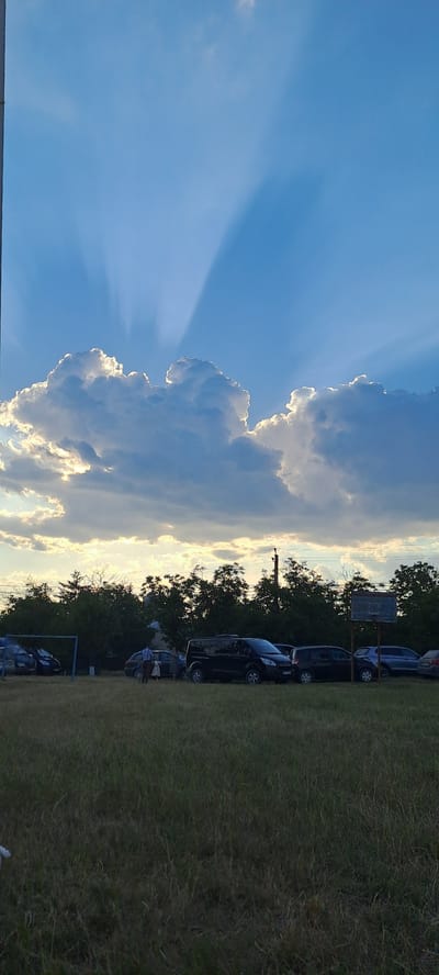 The image shows a sky with crepuscular rays emanating from behind a large cloud formation. Below the sky, there is a treeline. In front of the treeline, there is a grassy field with several parked vehicles and a few people. The scene appears to be an outdoor gathering or event. Based on the provided context, the image is located in Cotiujenii Mici, Moldova.