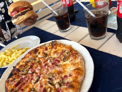 In Ravda, Bulgaria, a table is set for a meal with a hamburger being held, a pizza, french fries, and two glasses of cola with ice and straws. A bottle of cola is also visible, as is a menu partially obscured by the drinks. The meal appears to be served on a light-colored, slatted outdoor table covered with a dark blue fabric tablecloth. The background includes hints of a person's arm and clothing, suggesting a casual dining setting.