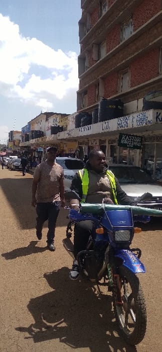 The image displays an urban street scene in Nyeri, Kenya, under a partly cloudy daytime sky. In the immediate foreground, an adult male with a dark complexion is seated on a blue dual-sport motorcycle. He wears a yellow reflective vest over a dark jacket, a tan collared shirt, black trousers, and white-and-black sneakers. His hands are on the handlebars, and his head is turned slightly right with a neutral to slight smile expression. The motorcycle has a prominent blue front fender with "SPORT" visible, a square headlight, and knobby tires.

To the left and slightly behind the motorcyclist, another adult male with a dark complexion walks away from the camera. He wears a patterned light brown shirt, dark trousers, dark shoes, and a dark baseball cap. The ground surface is an unpaved light brown dirt or gravel road, casting distinct shadows of both individuals and the motorcycle towards the upper left, indicating direct sunlight from the right.

Along the right side of the frame, a multi-story building, constructed of reddish-brown brick or tiles, ascends into the sky, revealing multiple window openings and a scaffold-like structure on one floor. Several black plastic water tanks are visible on an overhang of this building. In the mid-ground, along the left side of the street, a series of lower-rise commercial buildings feature storefronts with various signs, one partially legible as "KASTURI HOUSEHOLDS LTD." Further down the street, additional indistinct vehicles, including a white sedan, and pedestrians are visible.