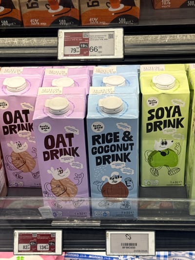 This is a medium shot photo taken inside a refrigerated display case at a grocery store in Odesa, Ukraine. The primary subjects are several cartons of plant-based milk drinks. From top to bottom, the visible drinks are Soya Drink, Rice & Coconut Drink, and Oat Drink, with multiple cartons of the Oat Drink stacked. The cartons feature colorful cartoon characters and text in English and Ukrainian, promoting their vegan status and lack of added sugar.
The scene is brightly lit, typical of a supermarket. On the right side of the image, a price tag holder is visible, displaying product information, prices in Ukrainian Hryvnia (грн), and expiry dates. One tag shows "24/06/2026" and "BF90C650". Other price tags show prices like "79 грн" and "66.99 грн". The expiration dates on the cartons are also visible, such as "10/03/27" and "12/11/26".
The focus is on the products themselves, suggesting a commercial or retail context. The overall impression is one of a well-stocked and organized food display. There are no people visible in the frame. The image is likely taken during daylight hours, given the internal lighting of the store.