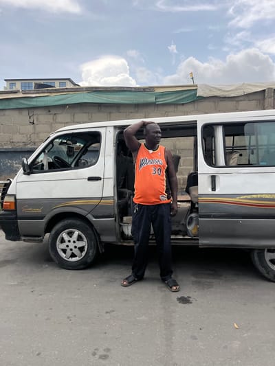 A man in an orange Golden State Warriors basketball jersey (#30) and black pants stands outside of a white and grey van. The man's left hand is on his head. He is wearing black sandals. The van is parked on an asphalt road. Behind the van, there is a cinder block wall with a green tarp partially covering it. A multi-story building with blue windows is visible behind the wall, and an antenna is visible on the roof of a structure farther back. The sky is cloudy. The location is Itirin, Nigeria.