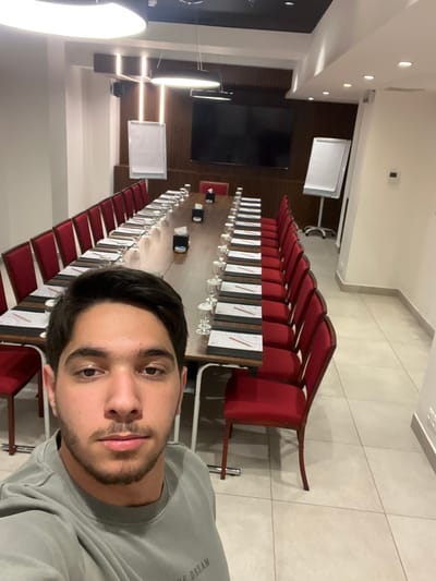 The image is a selfie taken by a young man in a large, formal conference or meeting room. The man, with dark hair, a beard, and a mustache, occupies the foreground on the right side of the frame, looking directly at the camera with a neutral expression. He is wearing a grey long-sleeved shirt with the text "LIVE DREAM" partially visible on his right shoulder.

The room is extensively set up for an event or meeting. A very long conference table extends from the mid-ground towards the back of the room, flanked by numerous red upholstered chairs on both sides. Each place setting on the table includes white papers or notebooks, pens, water glasses, and wine glasses, suggesting a formal event or dining arrangement.

The room's interior features light-colored tiled flooring and walls that combine light-toned panels with sections of dark wood paneling. Several whiteboards on stands are visible along the walls, and large black screens or monitors are mounted on the darker wood sections. The ceiling is well-lit with a combination of recessed spotlights and large, circular light fixtures. The room appears empty of other individuals, suggesting the photo was taken before or after a gathering. The setting is a professional conference facility in Al-Mazza Municipality, Syria.