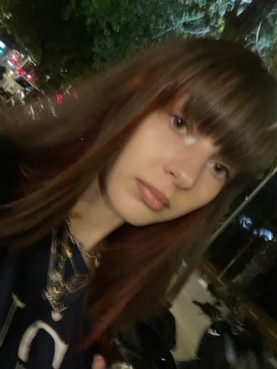 A young woman with long, reddish-brown hair and bangs poses for a close-up selfie. She has light skin, green eyes, and a neutral expression. She wears a dark t-shirt with white and yellow detailing. A chunky silver necklace is visible. The background appears to be outdoors at night, with blurry lights and foliage suggesting an urban setting. The overall image is slightly grainy, indicating low light conditions.