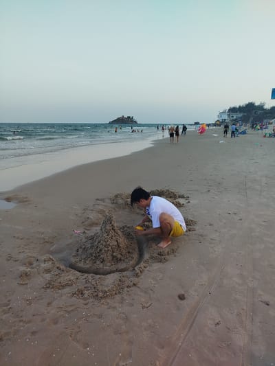 A young person is building a sandcastle on a beach. The person is wearing a white t-shirt and yellow shorts. They are squatting near a sandcastle, using a small yellow tool to mold the sand. The sandcastle is in the foreground. The beach stretches out in the background, with the ocean on the left. People are walking and swimming in the distance. A building is visible on the right side of the image. The sky is a pale blue. The image was taken in Ho Chi Minh City, Vietnam.