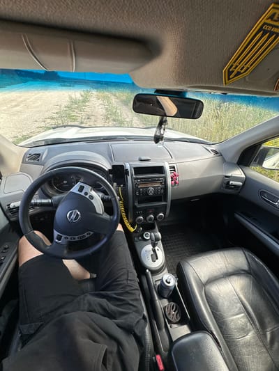 The image shows the interior of a car, viewed from the perspective of someone sitting in the driver's seat. The car's dashboard, steering wheel, gear shift, and part of the passenger seat are visible. A person's legs are also visible, wearing black shorts. The car appears to be parked outdoors, with sunlight streaming in through the windshield. The outside scene shows a sandy area with sparse vegetation, suggesting a rural or undeveloped location.

On the dashboard, there is a car stereo system, a mobile phone mount, and a yellow coiled wire. A rearview mirror is also visible, reflecting the interior of the car. On the passenger seat, a can of what appears to be soda is placed next to an air freshener. A black leather car seat is visible on the right side of the frame. There is a yellow emblem on the ceiling of the car.

The overall impression is a candid shot of a car interior during the daytime. The lighting suggests it is a sunny day. There are no explicit cues about the emotions of the person in the car, but the relaxed posture of the visible legs might suggest a casual setting. The image does not display any discernible text other than what might be on the car's branding or the can, which is not clearly legible. The location context provided is Abrykosivka, Ukraine, which aligns with the visual cues of a rural, outdoor setting.