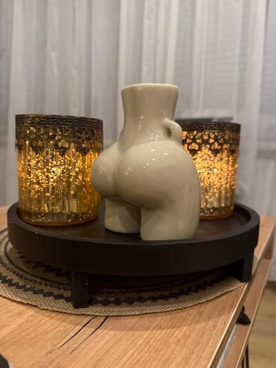 A beige ceramic statuette in the shape of a woman's buttocks is positioned on a dark wooden tray. The tray is placed on a light brown wooden table, with a woven circular placemat underneath. Flanking the statuette on the tray are two glass candle holders with ornate dark metal tops and a golden, textured glass base, both containing lit candles that emit a warm glow. The background consists of white sheer curtains. The scene appears to be indoors. The specific location in the city of Unknown, Russia is not discernible from the image.