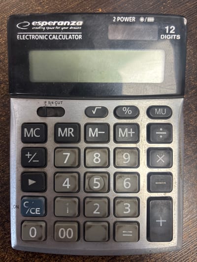 A black and silver Esperanza electronic calculator sits on a wooden surface. The calculator has a digital display screen and a keypad with numerical and functional buttons. The brand name "esperanza" is visible at the top left of the calculator, along with the words "ELECTRONIC CALCULATOR" below it. The top right of the calculator indicates "2 POWER" and "12 DIGITS". The wooden surface appears to be a table or desk with a visible wood grain texture. The image is well-lit, and the calculator is the primary subject. There are no people or other objects in the frame. The location is Līvāni, Latvia.