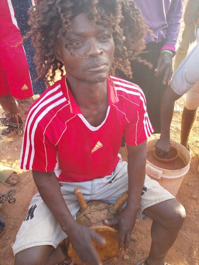 In Vodni, Nigeria, an outdoor scene depicts an adult male seated on reddish-brown ground, holding a traditional drum. The man has dark skin and prominent dark, curly hair. He wears a red short-sleeved polo shirt with white stripes on the shoulders and a gold Adidas logo on the right chest. His lower attire consists of light-colored shorts with a dark design on the left thigh. His hands are positioned on a light-brown, hide-covered drum with wooden pegs resting on his lap. Visible facial features include a mark or scar near his right eye.

Multiple individuals are partially visible around him. To the left, a person wears red shorts with an Adidas logo. Behind the main subject, another person in a purple top and dark trousers shows a hand with a green wristband. To the right, an individual's arm reaches into a white plastic bucket containing a reddish-brown substance and a smaller brown bowl. Footwear, including a light green sandal, is visible on the ground. The environment is characterized by dry, reddish-brown soil.