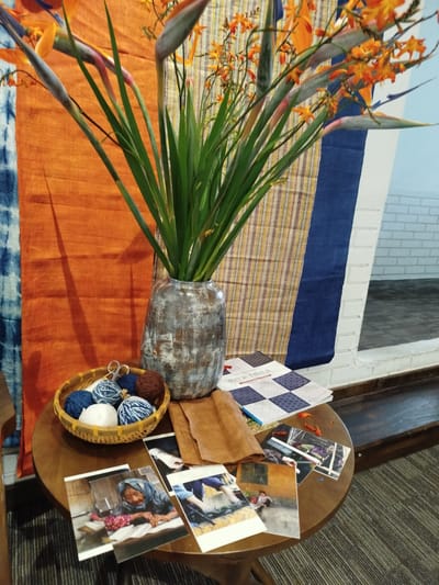 A still life scene is depicted on a circular wooden table, possibly in a workshop or cultural exhibition space. Dominating the background is a vibrant display of orange and blue textiles, creating a striking backdrop. To the left, a piece of indigo-dyed fabric with a tie-dye pattern is partially visible. The main textile is a bright orange, with a textured surface and a long, dark shadow cast across it. To the right of the orange fabric, another textile features vertical stripes in shades of yellow, red, and blue, with a thick navy blue band running down its right side.
In the center of the scene, a tall, grey, textured ceramic vase holds a generous bouquet of vibrant orange flowers and long, green leaves. The flowers appear to be Strelitzia, commonly known as bird of paradise, with their distinctive bird-like shape.
In front of the vase and to the left, a small, woven basket contains several balls of yarn in various shades of blue, white, and brown, along with a pair of small scissors.
Arranged on the table in front of the basket are several printed photographs, appearing to be mementos or part of an exhibit. One photograph prominently features an elderly woman wearing a blue headscarf, looking down at something on a table. Another shows a child sitting in front of a window. Other photographs show glimpses of people engaged in activities.
Also on the table, to the right of the yarn basket and in front of the flowers, are folded pieces of a light brown fabric, possibly linen or cotton. To the right of these, a book or brochure with a checkered pattern of dark blue and white squares is laid open.
The overall impression is one of craftsmanship and cultural display, with natural elements (flowers and yarn) combined with artisanal textiles and photographic documentation. The lighting suggests an indoor setting, possibly with natural light contributing to the warm tones of the scene. The location within Lâm Viên - Đà Lạt, Vietnam, is implied by the context of the request, suggesting a possible local craft or cultural presentation.