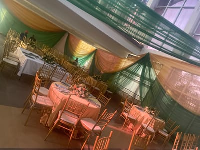 This media file shows an indoor event space decorated for a celebration. The scene is set with numerous gold chairs surrounding tables draped in peach-colored tablecloths. A floral arrangement of pink and white flowers is placed on one of the tables, suggesting a festive occasion such as a wedding or a formal party. The ceiling is adorned with drapes of green and peach fabric, creating a colorful and elegant ambiance. The lighting appears to be artificial, casting a warm glow over the setup, indicating the event is likely taking place in the evening or indoors with controlled lighting. The setting resembles a banquet hall or a reception area. There are no visible people actively participating in an activity, but reflections in the surfaces suggest a few individuals might be present or have been present in the space. The overall mood conveyed is one of celebration and elegance.