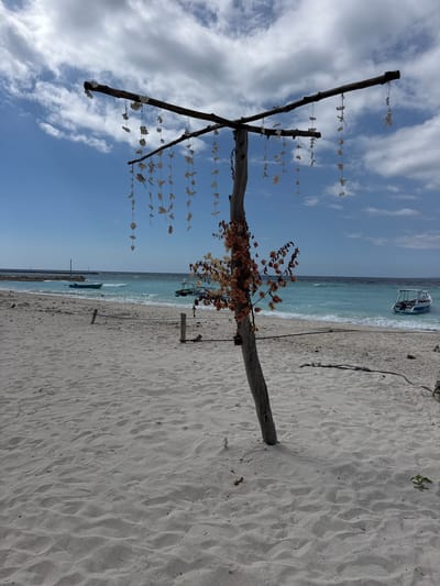 The image depicts a beach scene in an unspecified location in Indonesia. In the foreground, a weathered wooden post stands vertically, supporting a horizontal wooden beam adorned with strings of seashells. To the right of the post, a branch with dried, light-orange flowers is attached. The beach is covered in fine, light-colored sand with undulating patterns. In the middle ground, the turquoise ocean meets the shore with gentle waves. Several boats are visible on the water: a small green boat is closer to the shore, and a white boat with a blue trim is further out. In the background, a dark, calm sea stretches towards the horizon, meeting a hazy sky filled with white clouds against a blue backdrop. Wooden posts and ropes are strategically placed along the shoreline.