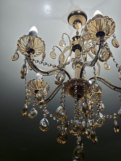 A crystal chandelier hangs from a white ceiling, illuminated by lights in the fixture. Ornate arms extend from the center, embellished with crystals in various shapes, suspended by delicate chains. The metal is a reflective silver or gold, and the ceiling transitions into a dark grey background to the right.
