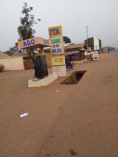 The image depicts a roadside scene in Ankpa, Nigeria, centered around a gas station and local businesses under an overcast sky. The road surface is a reddish-brown, possibly unpaved or gravel, and features a prominent, large, open rectangular drain or ditch in the midground, close to the viewer. A small piece of white litter lies on the road in the foreground.
To the left, a gas station is visible with a canopy displaying "AGO" in blue letters. Beneath it, a black fuel pump stands on a concrete base. Adjacent to this, a tall, yellow sign with "ITA" in blue at the top reads "AGO GLOBAL OIL-GAS NIG. LTD." in blue and green text. A smaller sign near the base of the main sign lists "PMS AGO Kerosene DPK". A tall, leafy tree provides a natural element behind the gas station structure.
In the midground, to the right of the gas station, a person on a blue and black motorcycle is partially visible, either parked or slowly moving along the road. Beyond the gas station and motorcycle, a series of simple buildings and market stalls with awnings line the road, suggesting a commercial area. Further down the road on the right, a white sign with "POS" is discernible.
The overall lighting is soft and diffused due to the overcast weather, indicating either early morning, late afternoon, or a heavily cloudy day. The scene conveys a functional, everyday atmosphere of a local service and business hub.
