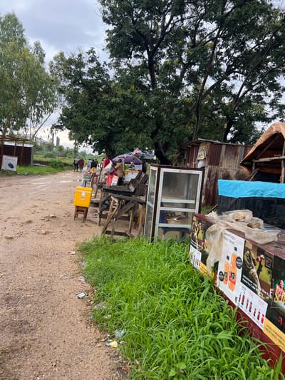 This image depicts an outdoor scene, likely a roadside market or informal settlement in Rafin Par, Nigeria. The foreground is dominated by a dirt path with scattered debris and a patch of lush green grass. To the right, a large display board advertising Coca-Cola products and other beverages is propped up. The board features vibrant images of bottles and cups, along with prices in Nigerian Naira (N).
Further into the scene, a makeshift stall is visible, constructed from corrugated iron and wood. Various items are displayed on tables, including what appear to be packaged goods and possibly food items. A prominent yellow cooler box sits on a stool in front of the stall. Several people are gathered around the stall, engaged in what seems to be commercial activity, though their faces are not clearly discernible. Some are standing, while others are seated. The atmosphere appears casual and busy, characteristic of a market day.
Behind the stall, a large, leafy tree provides shade. Further back, more trees and some distant buildings can be seen, suggesting a semi-urban or rural fringe setting. The sky is overcast, indicating a cloudy or possibly rainy day. The overall impression is one of everyday commerce and community life in a developing area.