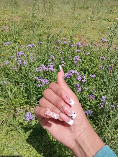 A close-up photograph captures a human right hand, exhibiting a medium skin tone, extended in an outdoor setting within Germiston, South Africa. The hand's thumb is raised, presenting a "thumbs up" gesture. All visible fingernails are long, artificial, and intricately adorned. The pinky nail features a light pink base with a white criss-cross pattern and a three-dimensional light pink bow. The ring finger nail has a white French tip with three small, clear rhinestones. The middle finger nail displays a white French tip accented by a black and white stylized animal face near the cuticle. The index finger nail also features a white French tip and a three-dimensional light pink bow. The thumb nail presents a plain white French tip. A teal-colored garment sleeve is partially visible at the bottom right. The background consists of dense green vegetation, including numerous clusters of small purple flowers and taller green leafy plants. A solitary yellow flower is visible on the left side of the midground. The scene is illuminated by direct sunlight, casting a distinct shadow of the hand onto the grass in the lower left foreground.