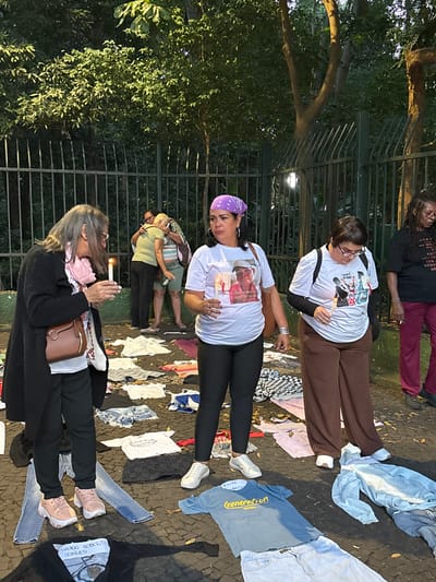 Several people are gathered in what appears to be a park or public space. They are standing amidst an array of clothing items laid out on the ground. Four individuals are distinctly visible. All the people are white except for one, a black woman. One woman is wearing a purple bandana, a white shirt with a photograph, black pants, and white tennis shoes, and she is holding a lit candle. Another person is wearing a white shirt with a graphic and brown pants. A third person is wearing a black overcoat and has a shoulder bag. The fourth individual is a black woman with burgundy pants and is wearing a black shirt with printed text. Some of the clothing items on the ground have writing or labels, including one with the words "Generation Brasil" and one with "THIAGO ROBERTO SOARES". The overall impression suggests a somber gathering or vigil.