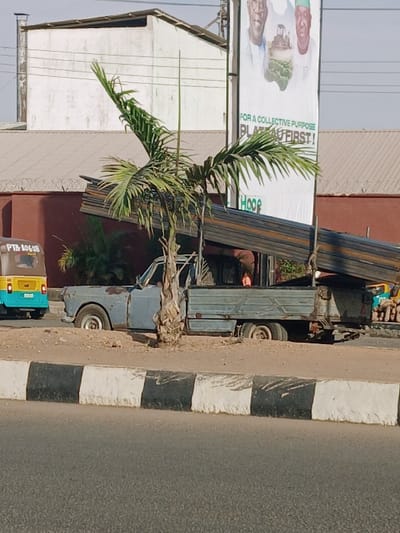 The image displays an outdoor scene, likely a street or roadside area, under bright daylight. The primary focus is on a parked large truck, a palm tree, and a billboard, with other vehicles and buildings in the background.
In the foreground, on the right side of the image, is a segment of an asphalt road. Adjacent to the road is a curb or barrier painted with alternating black and white stripes. To the left of the curb, on an unpaved shoulder, several vehicles are parked.
A large, dark-colored truck, possibly a flatbed or open-top cargo truck, is parked facing right, with its rear towards the viewer. It appears to be laden with long, dark, possibly metallic or wooden beams or pipes. A person is visible in the driver's cabin of this truck. Behind this larger truck, and partially obscured by a palm tree, is a smaller, older-model light blue or grey truck. Further to the left, at the bottom of the image, a portion of a yellow and blue three-wheeled vehicle, resembling an auto-rickshaw or tuk-tuk, is visible. The license plate "PTN-10620" is discernible on this vehicle.
A medium-sized palm tree with green fronds stands prominently in the center-left, between the parked trucks and the background structures. Its foliage partially covers the smaller truck and extends upwards, past the billboard.
Behind the palm tree and trucks, a large white billboard is visible. The billboard features images of people or figures and some text. Visible text includes "FOR A COLLECTIVE PURPOSE" and "PLANT FIRST!", with the word "PLANT" partially obscured but clear. Below this text, the word "Hoop" is also visible, possibly a brand or organization name.
In the background, behind the billboard and trucks, are several buildings. One building on the left has a light-colored exterior with horizontal siding. Another building behind the trucks appears to have a reddish-brown facade.
The overall impression is a bustling roadside scene, characteristic of an urban or semi-urban environment in a warm climate. The clear sky suggests fair weather.