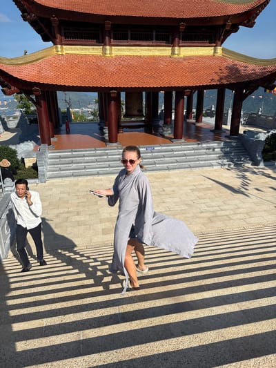A woman in a grey robe and sunglasses holds a phone while walking on a paved surface. She is facing the camera. A man in a white shirt and black pants is lying on the same surface, talking on a cell phone. A traditional building with a red and gold roof is visible on the left. The environment suggests a location in or near the city of Diên Khánh, Vietnam, based on the architecture and landscape. Sunlight creates strong shadow patterns on the ground.