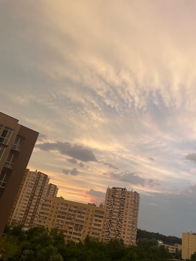 The image shows a view of buildings and the sky in Brovary, Ukraine. The sky is filled with dramatic clouds, tinged with the warm colors of sunset or sunrise, ranging from pale yellow to soft pink and gray. Streaks of cirrus clouds are visible, creating a wispy texture across the sky.
In the foreground, a portion of a modern building with a beige facade and numerous windows is visible. To the right and slightly further back, two taller apartment buildings dominate the scene. The first of these has a distinctive black and white patterned facade, resembling animal print or a stylized wave. The second, behind it, is a more standard, light-colored residential building with many windows. Both buildings appear to be multi-story, typical of urban residential architecture.
To the far right and in the background, a dense line of dark green trees runs along a hillside, suggesting a natural landscape adjacent to the urban development. A sliver of another building, taller and lighter in color, can be seen at the very top right edge of the frame.
The overall impression is one of a serene evening or morning scene in a residential area. The dramatic sky suggests a time of day with soft, diffused light, likely around sunset or sunrise. There is no visible text in the image. The composition, with buildings angled against the expansive sky, creates a sense of depth and perspective.