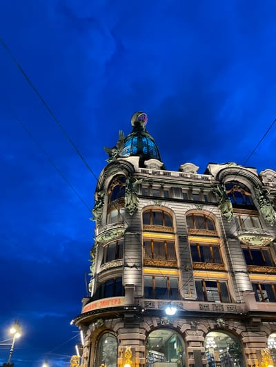 The image captures an architectural detail of the Singer House in Saint Petersburg, Russia, under a dark blue sky with visible clouds and power lines. The building features ornate Art Nouveau design, with a prominent domed roof structure illuminated by blue and purple lights. Decorative elements, including winged figures and intricate carvings, adorn the facade. The lower levels of the building display arched windows and the inscription "КОМПАНИЯ ЗИНГЕРЪ" in red lettering. Streetlights and interior lights of the building are visible, suggesting it is evening or night. The scene is captured from a low angle, emphasizing the grandeur of the architecture against the night sky.