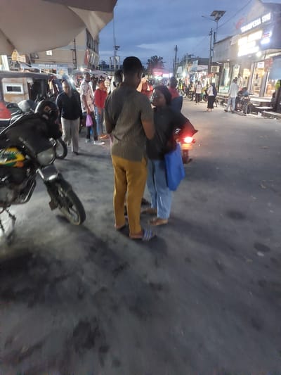 The image depicts a street scene at dusk in Dar es-Salaam, Tanzania. Two individuals stand near a motorcycle parked on the street. One person is wearing a striped shirt and mustard-colored pants, while the other is wearing a dark shirt and light-colored pants. The motorcycle is black with a round headlight. A crowd of people and shops line the street, illuminated by artificial lights. The sky is a gradient of blues and grays, indicating twilight.