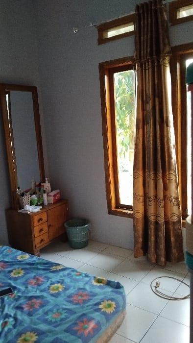 The image shows the interior of a bedroom in a home located in Panjalu, Indonesia, at coordinates -7.142 latitude and 108.278 longitude. The room has light blue walls, a wooden floor, and a white tiled floor. There is a bed with a blue, yellow, and pink floral patterned bedspread. In the background, there is a window with a wooden frame and a brown patterned curtain. To the left of the window, there is a dresser with three drawers and a mirror above it. The dresser is filled with assorted items. There is a blue plastic wastebasket to the right of the dresser. An electrical cord runs across the white tiled floor, winding through a hole in the floor. The bed is unmade. No people are present. The lighting is dim, possibly from the window which provides natural light. The scene appears to be a typical bedroom, unkept with personal items scattered throughout.