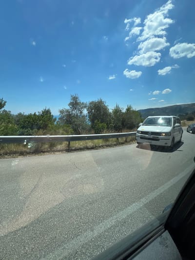 The image depicts a view from a moving vehicle on a road in Mattinata, Italy, on a sunny day with a bright blue sky and scattered white clouds. The road curves to the left, bordered by a metal guardrail on the left side. Beyond the guardrail, there is lush green vegetation and trees, with a glimpse of the sea in the distance.
In the foreground, the asphalt road is visible, with a white lane marking. A white Volkswagen Transporter van is driving on the road in the same direction as the camera. The sun is reflecting brightly off the windshield of the van. To the right of the van, another dark-colored car is partially visible. The overall scene is one of travel through a scenic coastal landscape.