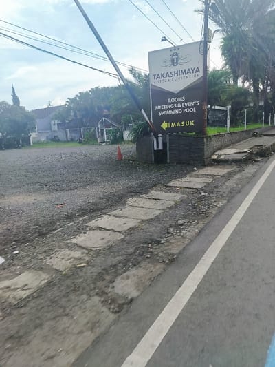 The image shows a sign for the Takashima Hotel and Convention located in Lembang, Indonesia at the coordinates -6.816 latitude and 107.619 longitude. The sign is positioned on a low stone wall bordering a gravel parking lot. It reads "Takashima Hotel and Convention" in white text on a brown background with four stars above. Below the name, a brown sign with white text states "Rooms, Meetings & Events, Swimming Pool" and an arrow pointing right with the word "Masuk" (Indonesian for "Entrance") beneath. The sign is situated in a grassy area with a large tree in the background. The sign is set on a low stone wall with a paved area on the left side of the image leading to the wall. The parking lot is empty except for a small orange cone on the left side of the image. The wall is partially cracked and eroded in areas. A paved road with a white painted line runs diagonally across the bottom of the image, cutting through the gravel parking lot on the left. The road is partially covered with debris. The environment is slightly overgrown with grass and weeds, suggesting the area is not frequently used. The image is taken from the perspective of a person standing on the road looking at the sign. The image was taken during the day. The sky is overcast with a few clouds. The sign is the only subject in focus. The parking lot and the road are out of focus.