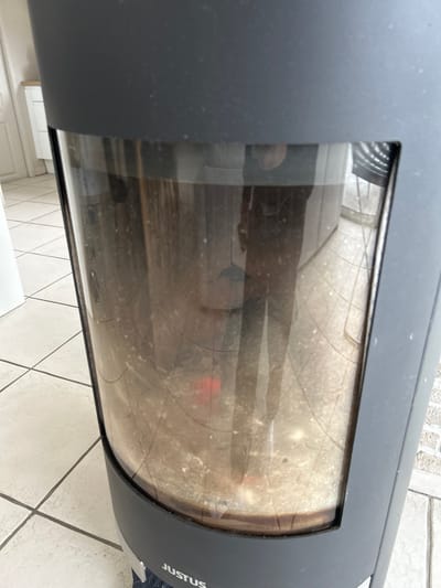 An interior scene captured in Veauchette, France, depicts a dark grey, modern, cylindrical "JUSTUS" brand wood-burning stove positioned on a light-colored tiled floor with grey grout lines. The stove's prominent glass viewing panel is significantly smudged and dusty. Through the soiled glass, a residual layer of ash and partially combusted material is visible within the combustion chamber, with a faint reddish glow suggesting the presence of embers or residual heat at the lower-middle portion. A distinct reflection of a person, identifiable by dark legwear and a lighter torso, is superimposed on the stove's glass. The reflection also shows more of the tiled floor extending away from the stove and a white cabinet or piece of furniture in the background to the left.
