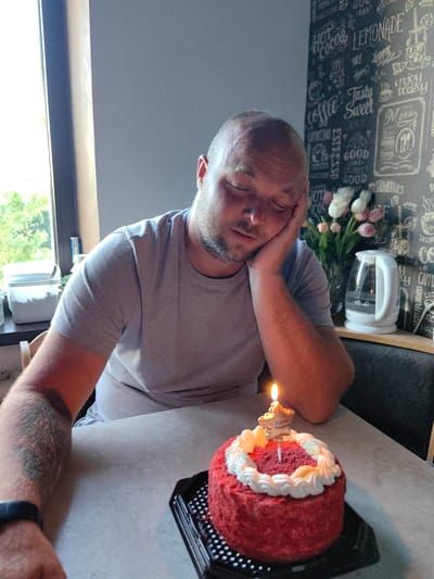 In this medium shot, a man with closed eyes and his head resting on his hand sits at a table. He appears to be napping, with his birthday cake, adorned with a lit candle and a star-shaped topper reading "С Днем Рождения" (Happy Birthday), in front of him. The cake is red velvet with white frosting. The man wears a grey t-shirt and a smartwatch. A tattoo is visible on his left arm.
The background features a wall decorated with a chalkboard-style print of various coffee and dessert-related words like "HOT COCOA," "LEMONADE," "COFFEE," "CAPPUCCINO," "AMERICANO," and "STRAWBERRIES." A bouquet of pink and white flowers sits in a vase, next to a white electric kettle. A window with greenery visible outside is to the left of the frame, suggesting an indoor setting. The lighting indicates it's daytime. The overall mood is quiet and perhaps a little melancholic, given the man's apparent sleepiness on his birthday. The location is identified as Fanipal, Belarus.