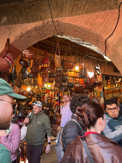 An indoor market scene within a souk in Marrakesh, Morocco, is depicted under a prominent, rough-textured, reddish-brown archway, likely constructed from plastered brick. Exposed black electrical wires run along the arch structure. A corrugated metal sheet is visible above the merchandise, serving as a lower ceiling or covering.
Hundreds of leather bags and purses of various sizes, shapes, and shades of brown, including shoulder bags, cross-body bags, duffel bags, and smaller pouches, are densely suspended from metal rods and hooks, dominating the visual space. Some bags feature fringe, intricate tooling, or simple designs. Multiple warm-toned incandescent light bulbs hang among the displays, illuminating the goods. In the background, additional merchandise includes colorful leather slippers (babouches) and decorative metal objects.
Approximately eight individuals, appearing to be patrons, are present in the crowded aisle.
* In the far foreground, partially visible on the left, is a bearded individual wearing a green baseball cap and glasses, facing right.
* To their right, a person in a pink top is partially visible.
* To the left of center, a male with a short beard, wearing a grey/olive green hooded sweatshirt, dark trousers, and a white flat cap with glasses on top, holds a white cylindrical object (possibly a vape pen) in his right hand. A dark smartwatch is on his left wrist. He looks left.
* Behind him, a male with short hair and glasses wears a light blue/purple collared shirt, gazing upwards and to the right.
* Further right, facing away from the camera, a person with dark, curly hair wears a dark padded jacket and a backpack.
* To their right, also facing away, a female with dark hair in a bun wears a brown leather jacket with a red collar visible underneath.
* On the far right, a male with dark curly hair and glasses wears a grey hooded sweatshirt with partial white lettering, looking left with his mouth slightly open, tongue partially extended.
* In the background on the left, an older male with grey hair and glasses, wearing a brown jacket, stands near a display, holding an object in his right hand and looking right.
* A partially visible individual with light hair is in the far right background.
The scene portrays a busy shopping environment.