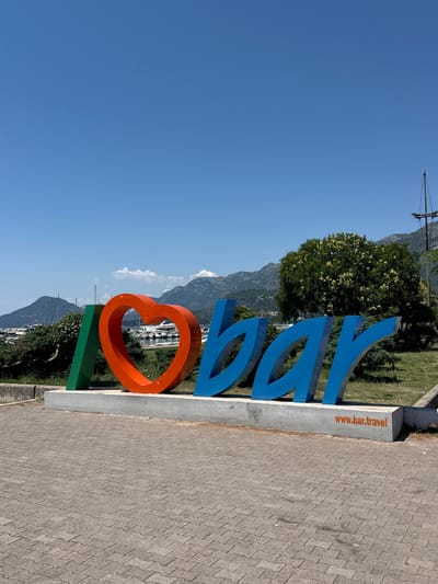 A large, colorful, 3D sign spelling out "I LOVE BAR" is displayed outdoors. The "I" is represented by a green rectangle. The "LOVE" is depicted by a large, orange heart. The letters "BAR" are rendered in blue with a distinctive, stylized font. The sign is situated on a gray concrete base, with the website "www.bar.travel" printed in orange on the right side of the base. The background reveals a scenic view of a bay dotted with sailboats and yachts, lush green trees, and rolling mountains under a clear blue sky. The foreground is paved with gray interlocking bricks.