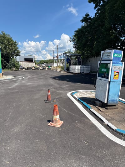 The image depicts an outdoor scene at what appears to be a small, possibly informal, service station or car wash facility in Ihtiman, Bulgaria. The foreground is dominated by a freshly paved asphalt surface with white and blue painted markings along the edge of a driveable area. Two orange traffic cones, one closer and one further away, are placed on the asphalt.

In the middle ground, to the right, stands a blue and white diesel pump. It has a display screen showing "4" and a sticker with "DIESEL" and a yellow warning sign with "ATTENTION" and "ANGLES MORTS" in French. To the left of the pump, there are several large white plastic tanks (likely for water or other liquids) and some stacked tires against a concrete block wall. In the background, a light-colored building with an open-fronted section is visible, with more tires stacked in front of it. Beyond the building, trees and distant hills are seen under a bright blue sky with scattered white clouds.

The overall impression is of a functional, albeit somewhat utilitarian, service area, possibly in a more rural or industrial part of Ihtiman. There are no people visible in the frame.